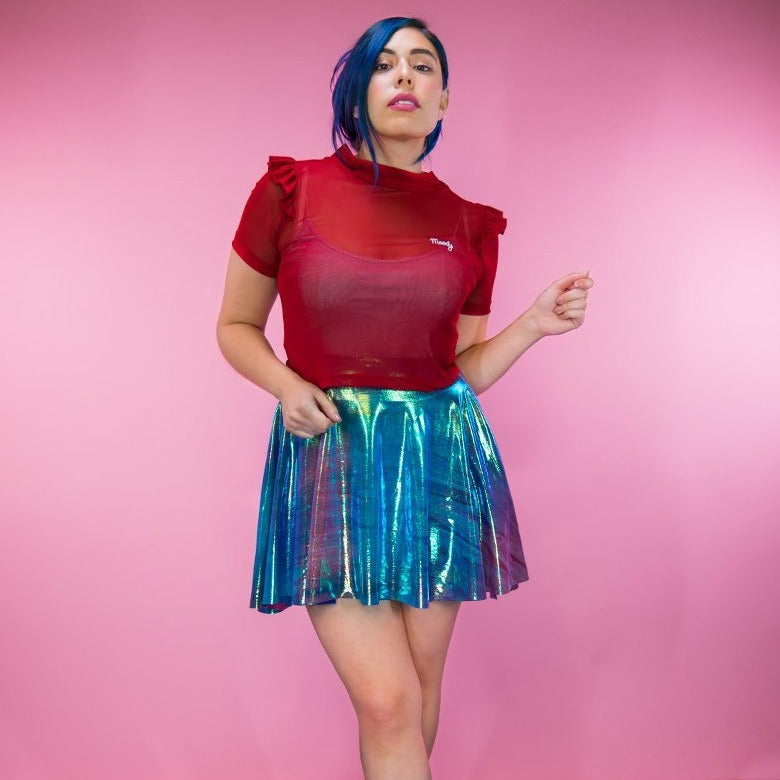 A woman wearing a bright red semi-sheer top and a shimmering holographic pleated skirt stands against a pink background. She has blue hair and is looking to the side with a neutral expression.